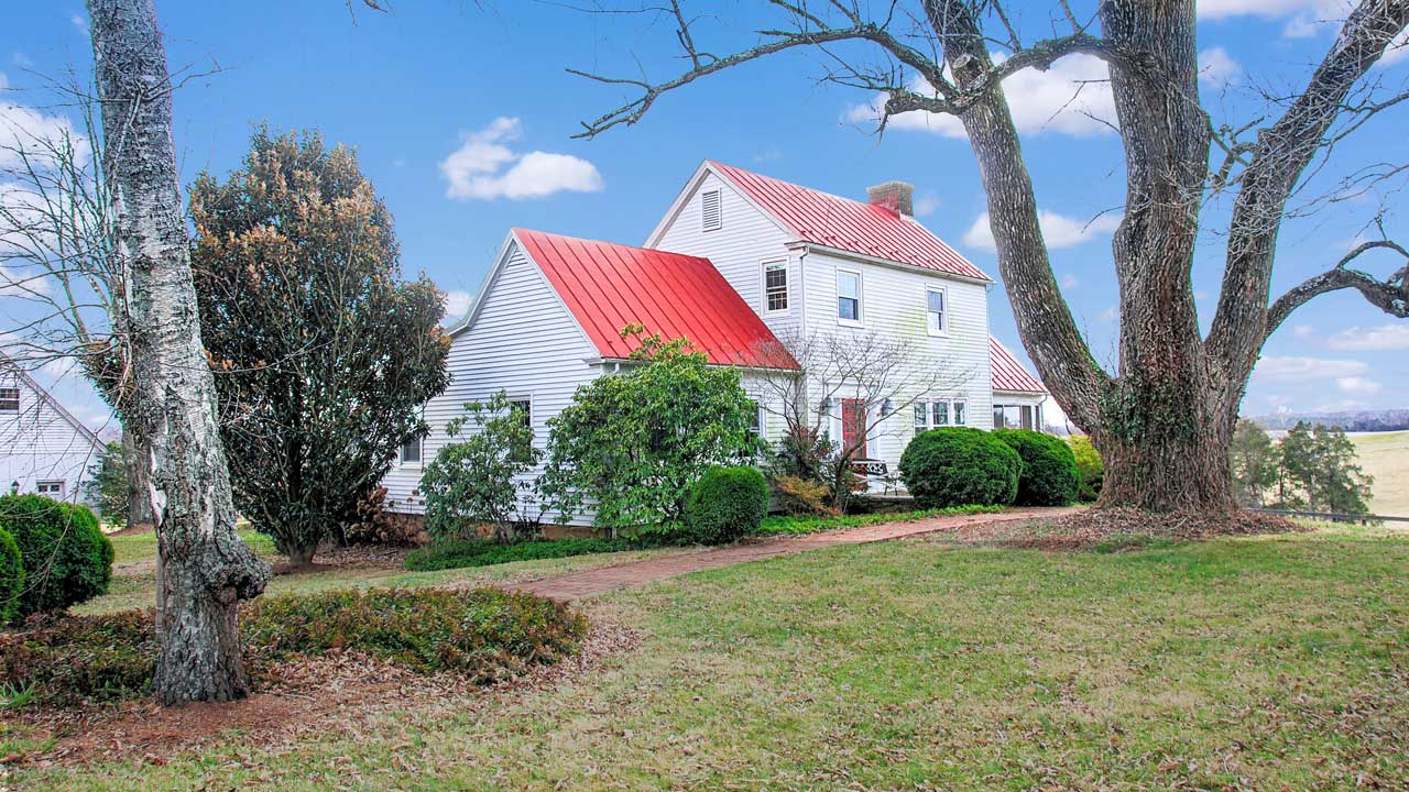 A charming historic farmhouse with white siding and a striking red metal roof, nestled among lush greenery. The home features a traditional design with a central chimney, multiple gables, and a welcoming red front door. A brick pathway leads to the entrance, framed by neatly trimmed bushes and mature trees, including a large, weathered tree in the foreground. The property sits under a bright blue sky with scattered clouds, evoking a classic country aesthetic.