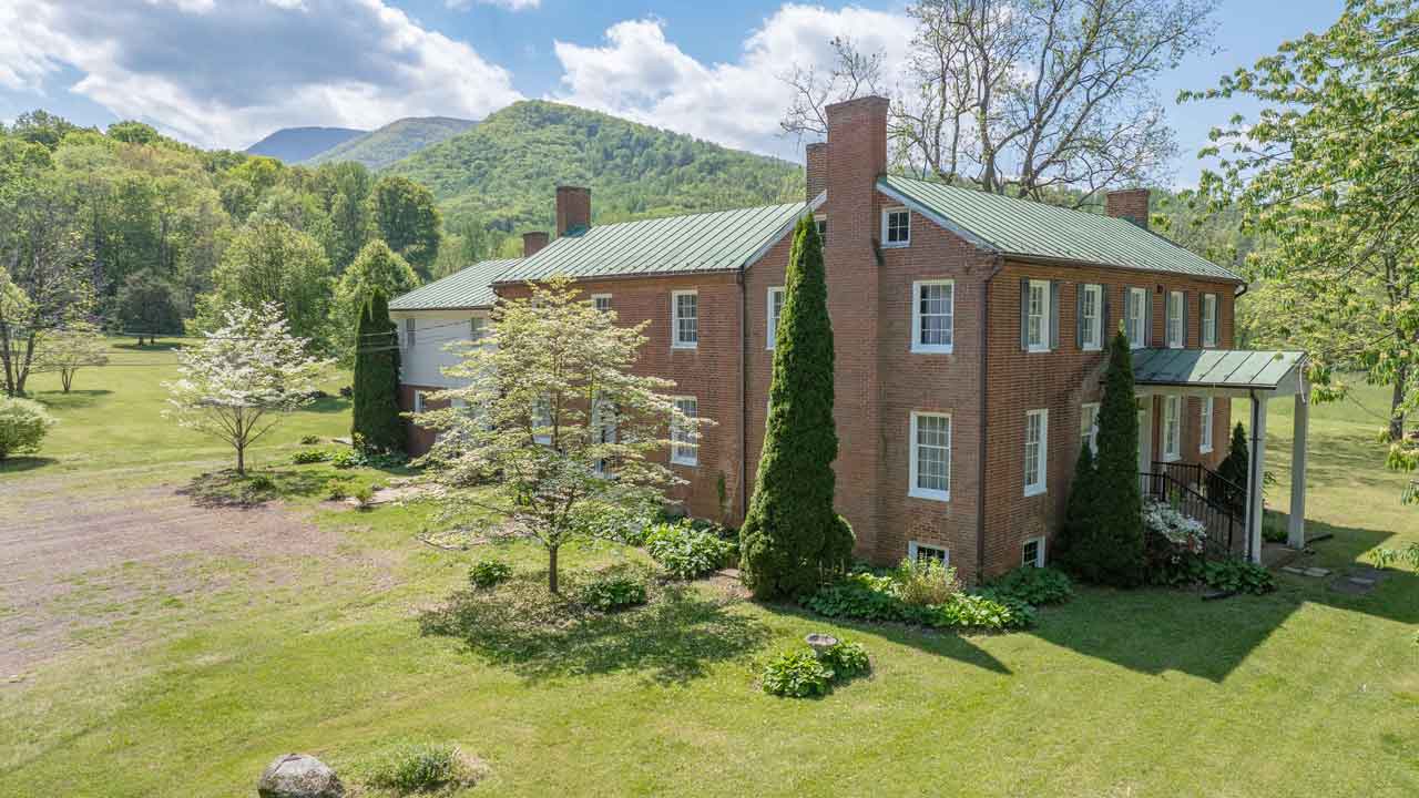 A historic two-story brick home with a green metal roof, multiple chimneys, and a classic symmetrical facade. The house features tall windows with white trim and a covered side entrance with a small portico. Lush landscaping surrounds the property, including mature trees, shrubs, and flowering plants. Rolling green hills and mountains provide a scenic backdrop under a bright blue sky with scattered clouds.