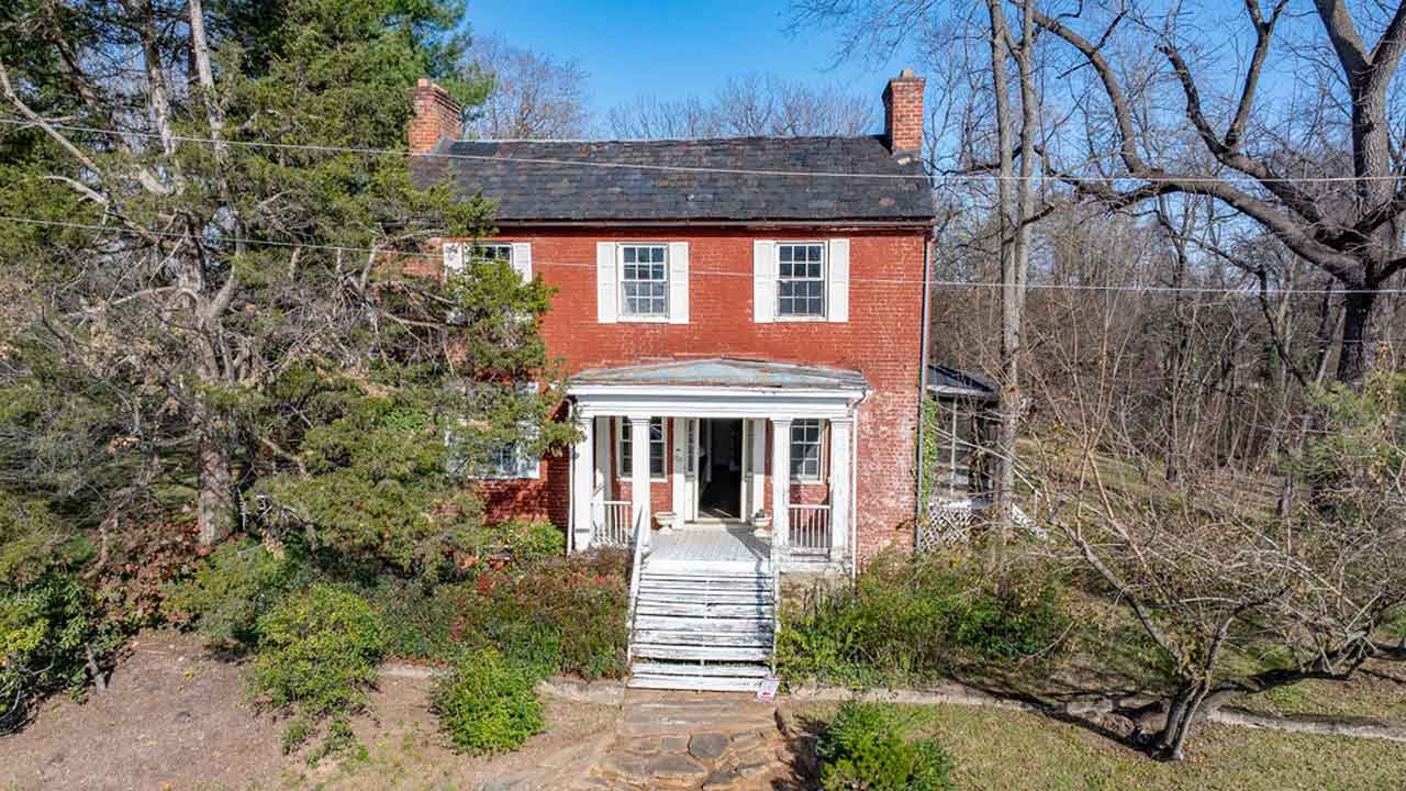 A historic two-story red brick home with white shutters and a weathered front porch, featuring a set of wooden stairs leading to the entrance. The home has a symmetrical facade with a slate roof and two brick chimneys. Overgrown shrubs and mature trees surround the property, hinting at its aged charm and renovation potential. The front door is open, revealing a glimpse of the interior.