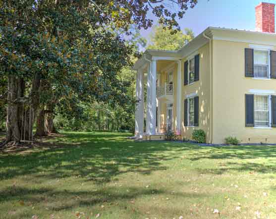 A stately two-story Southern-style home with a soft yellow exterior, black shutters, and a white columned portico, set amidst a lush green lawn and framed by mature trees. Sunlight filters through the foliage, casting dappled shadows on the grass, enhancing the home's timeless elegance and tranquil setting.
