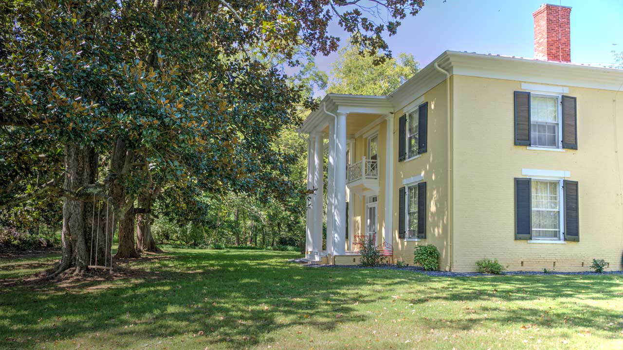 A stately two-story Southern-style home with a pale yellow brick exterior, black shutters, and a grand columned portico. The entrance features a second-story balcony with a decorative railing, and a red brick chimney rises above the roofline. The house is surrounded by lush green lawn and mature trees, creating a serene and elegant setting.