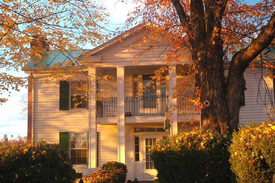 A historic two-story Southern-style home with white siding, green shutters, and a metal roof, bathed in warm golden light. The home features a grand front porch with white columns and a second-story balcony with decorative wrought iron railings. A large tree with autumn foliage partially frames the house, and manicured bushes surround the entrance.