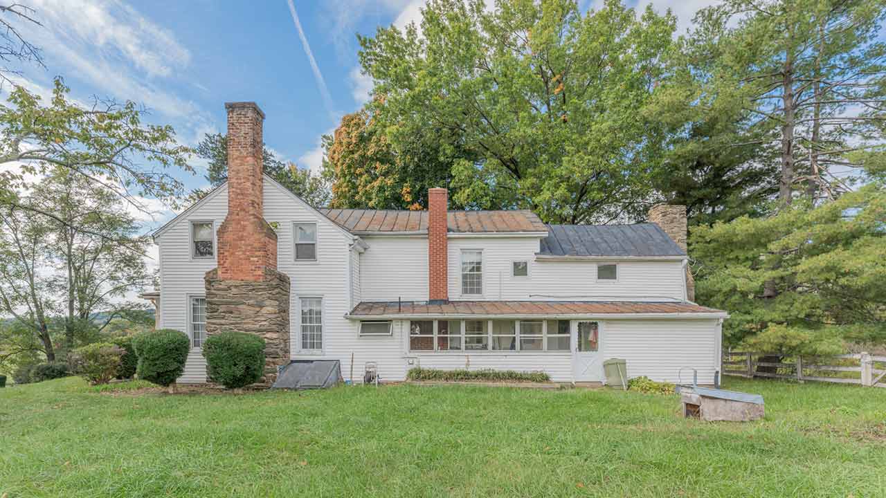 A rustic farmhouse with white siding, multiple chimneys, and a mix of metal and shingle roofing, set against a backdrop of mature trees and a blue sky. The home features a large stone chimney on one side, a long enclosed porch, and a basement entry with cellar doors. The surrounding lawn is lush and green, adding to the property's peaceful rural charm.