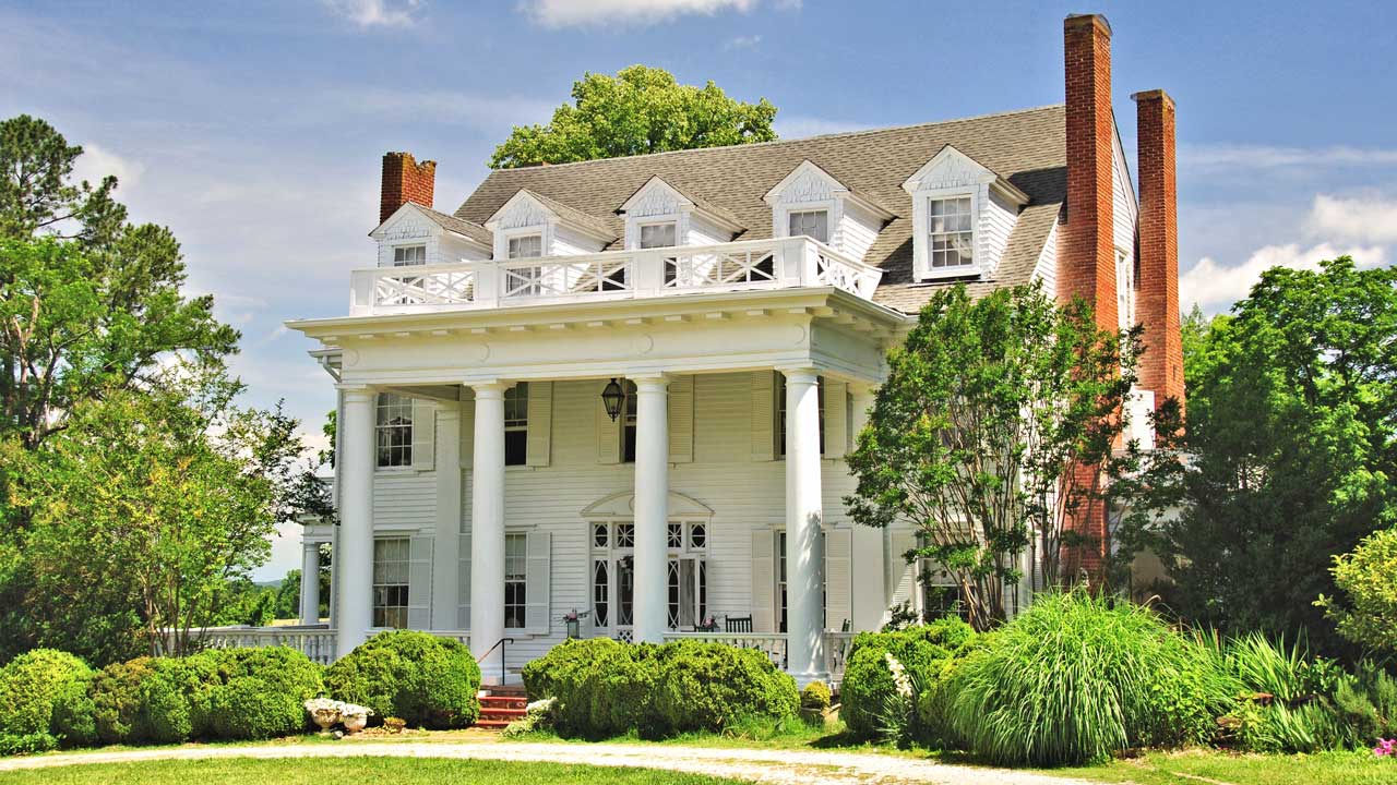 A grand Southern-style mansion with white siding, a symmetrical facade, and a large columned portico. The home features multiple dormer windows, a second-story balcony with a decorative railing, and two tall brick chimneys. Lush greenery and manicured bushes frame the entrance, while rocking chairs on the porch add to its inviting charm. A gravel driveway leads up to the historic estate under a bright blue sky.