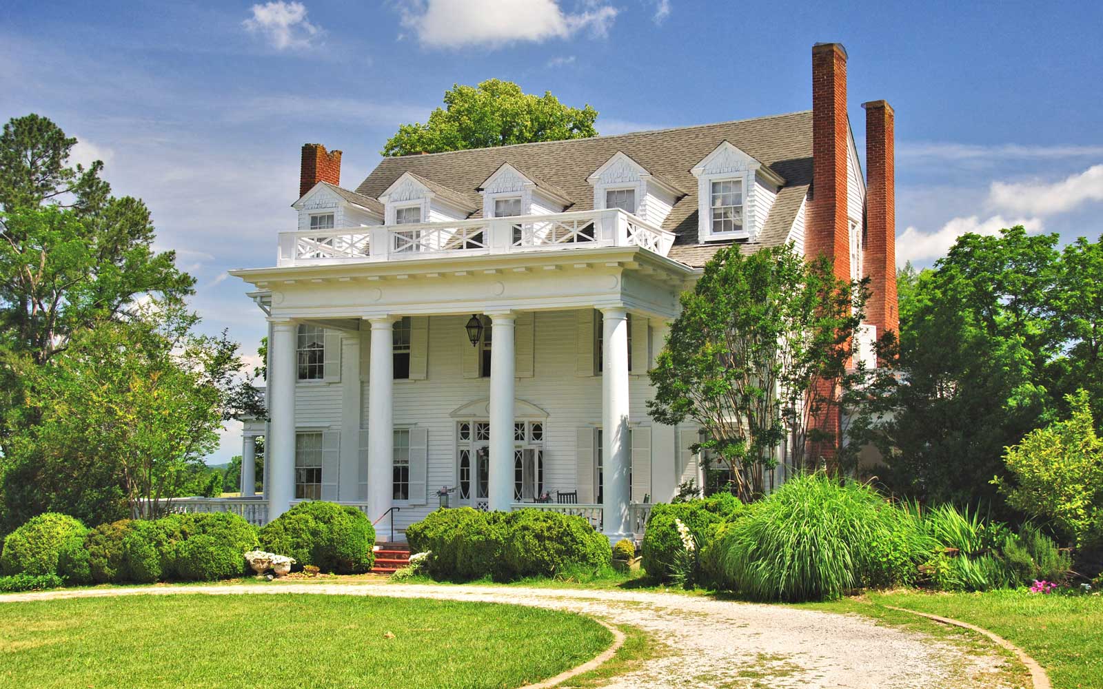 A grand Southern-style mansion with white siding, a symmetrical facade, and a large columned portico. The home features multiple dormer windows, a second-story balcony with a decorative railing, and two tall brick chimneys. Lush greenery and manicured bushes frame the entrance, while rocking chairs on the porch add to its inviting charm. A gravel driveway leads up to the historic estate under a bright blue sky.