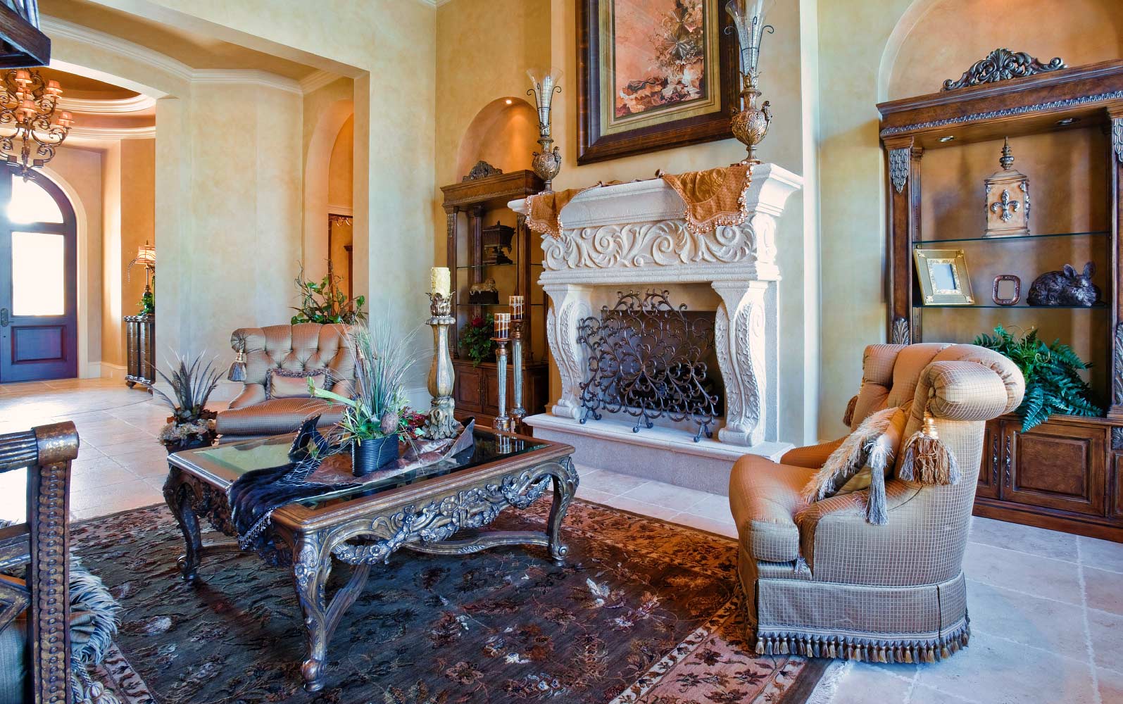 A luxurious living room featuring an ornate carved stone fireplace with intricate detailing, draped with decorative fabric. The space is adorned with elegant furnishings, including a richly carved wooden coffee table, plush upholstered seating, and tall candlesticks. A framed painting hangs above the fireplace, and a wooden display cabinet with glass doors stands in the background, enhancing the room’s sophisticated ambiance.