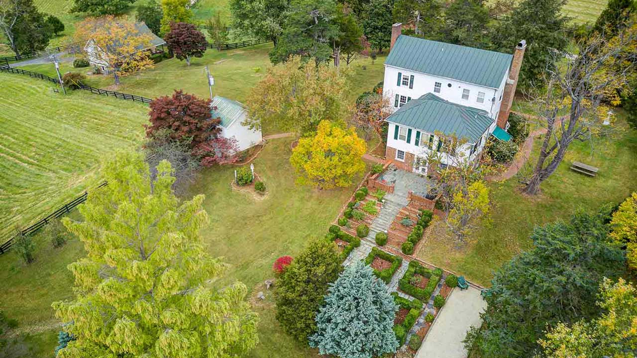 Aerial view of a historic two-story white farmhouse with a green metal roof, nestled among mature trees and surrounded by a landscaped garden. A symmetrical brick and stone walkway leads to the entrance, flanked by neatly trimmed hedges and topiary. The property features multiple brick chimneys, an adjacent outbuilding, and expansive green lawns with scattered trees and a wooden bench.