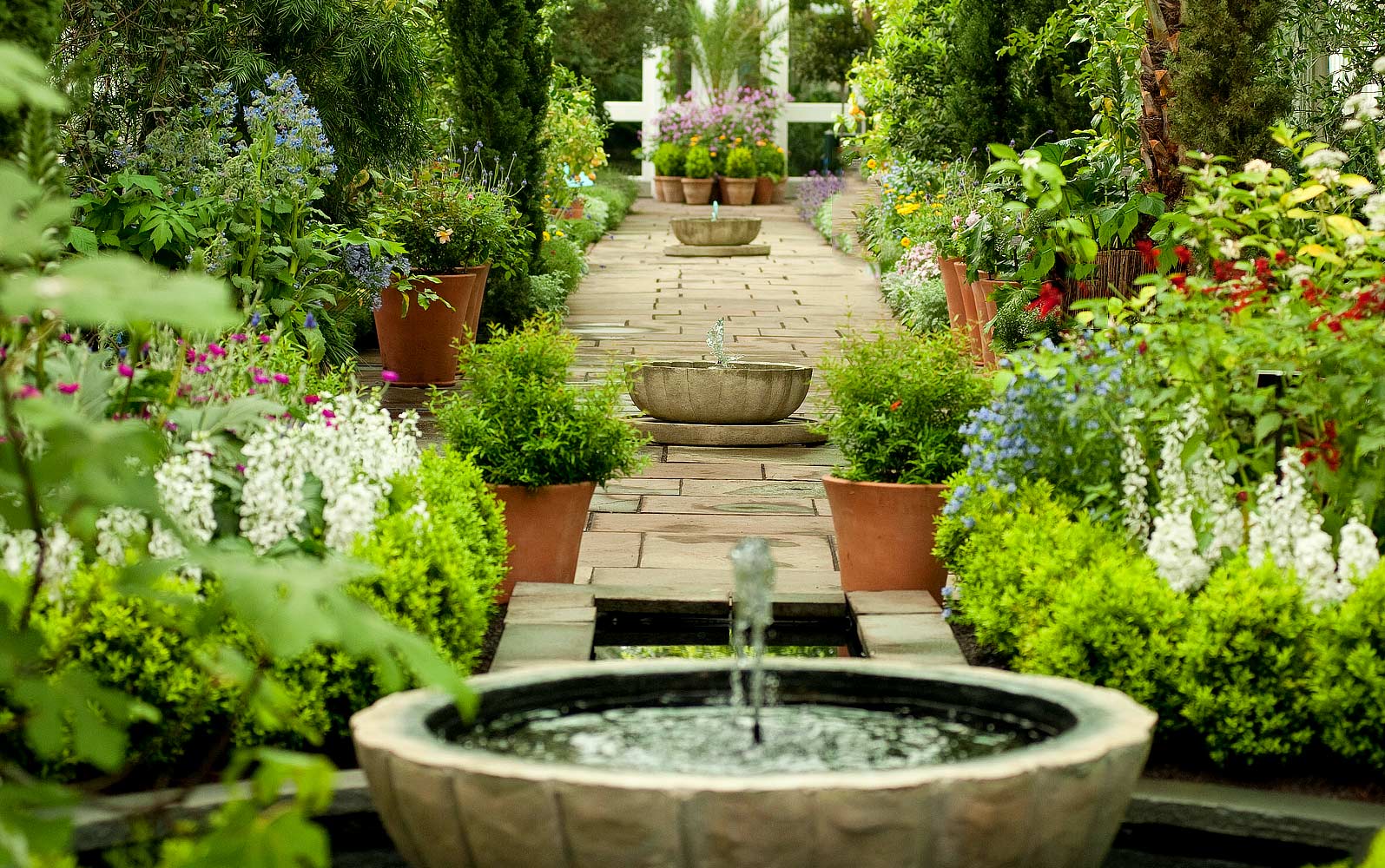 A beautifully landscaped garden pathway with a series of tiered stone fountains, surrounded by lush greenery and vibrant flowers in terracotta pots. The stone walkway is lined with neatly trimmed boxwoods and colorful blooms, leading to a white trellis adorned with more potted plants in the background. The tranquil water features create a serene and inviting atmosphere.