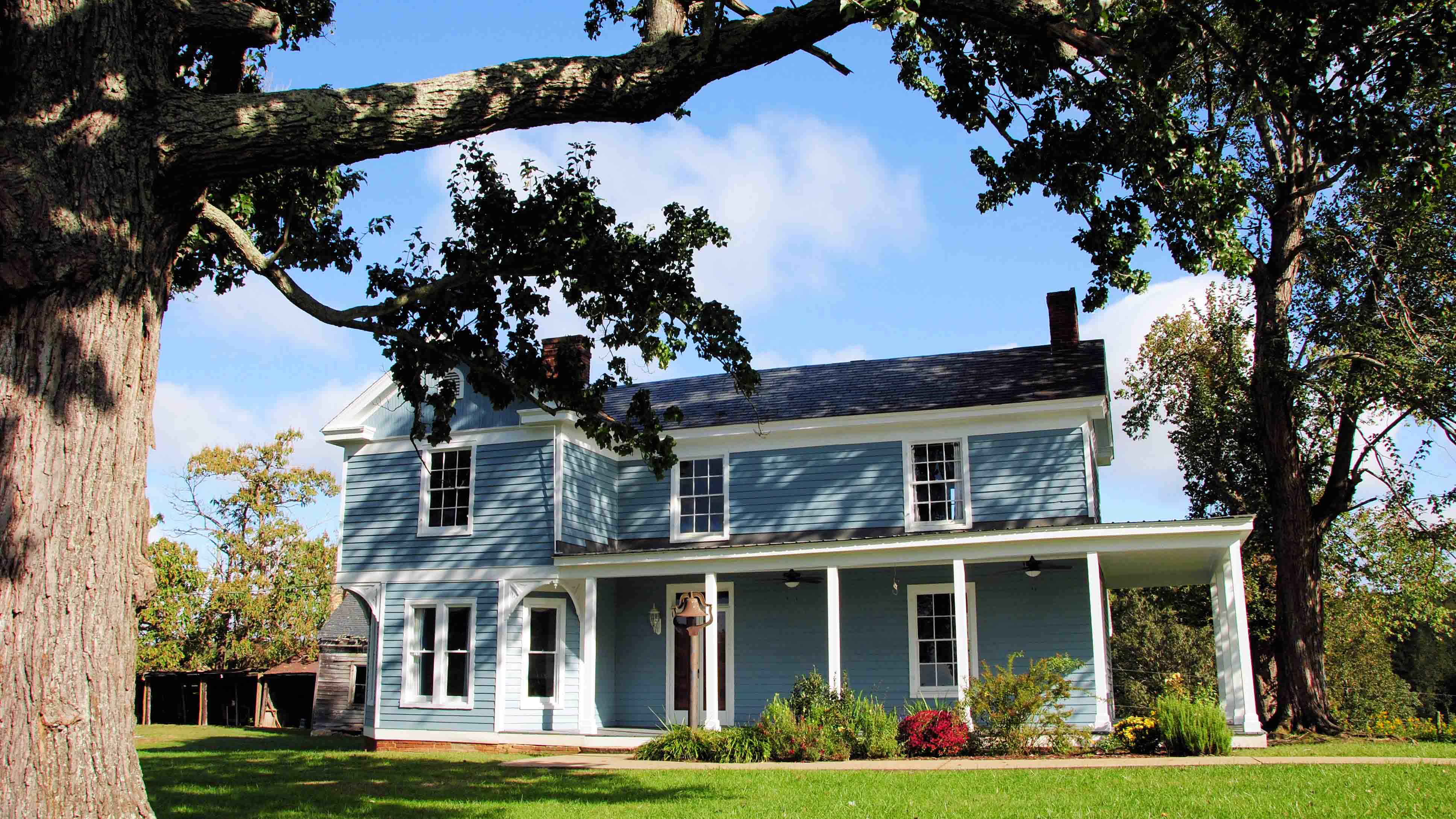 A picturesque two-story farmhouse with a blue exterior, white trim, and a welcoming wraparound porch. The home features a classic gabled roof, brick chimneys, and large windows that enhance its historic charm. Surrounded by lush green lawns, mature trees, and colorful landscaping, the house sits under a bright blue sky with scattered clouds, evoking a serene countryside setting.