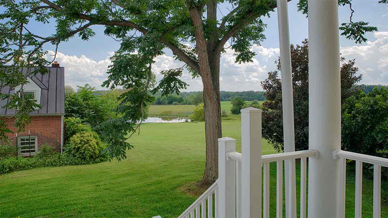 A scenic countryside view featuring a lush green lawn, a mature tree with sprawling branches, and a tranquil pond in the distance. To the left, a portion of a historic red brick house with a metal roof and dormer window is partially visible, surrounded by dense foliage. A white railing in the foreground suggests the vantage point is from a porch or deck, overlooking the peaceful rural landscape under a partly cloudy sky.