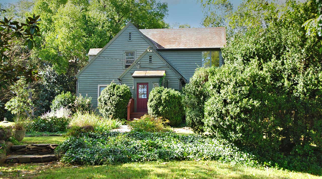 A charming cottage-style home with muted blue-green siding, a steeply pitched roof, and a welcoming red front door. The house is surrounded by lush greenery, including neatly trimmed bushes, ivy-covered ground, and mature trees that provide a secluded feel. A small wooden porch with a gabled entry adds to the home's character, while dappled sunlight filters through the trees, enhancing the peaceful atmosphere.